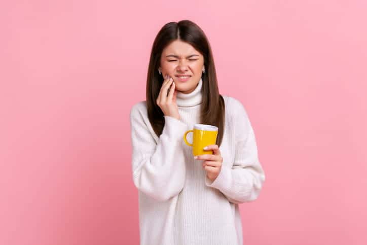 A woman holding a coffee cup while also holding her face in pain due to a dental emergency. 